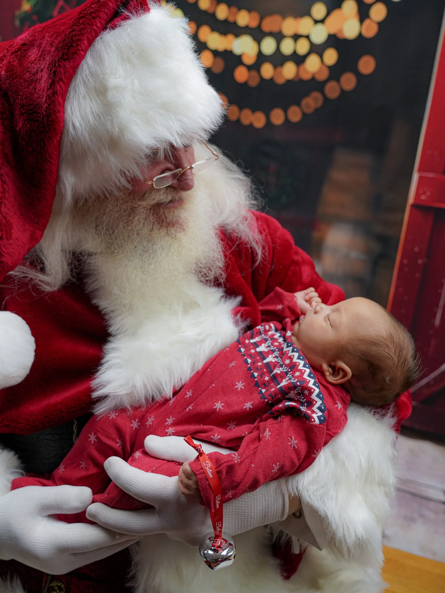 Santa Claus in a red suit holding a sleeping baby dressed in a red Christmas outfit with mittens, in front of a decorated Christmas background.