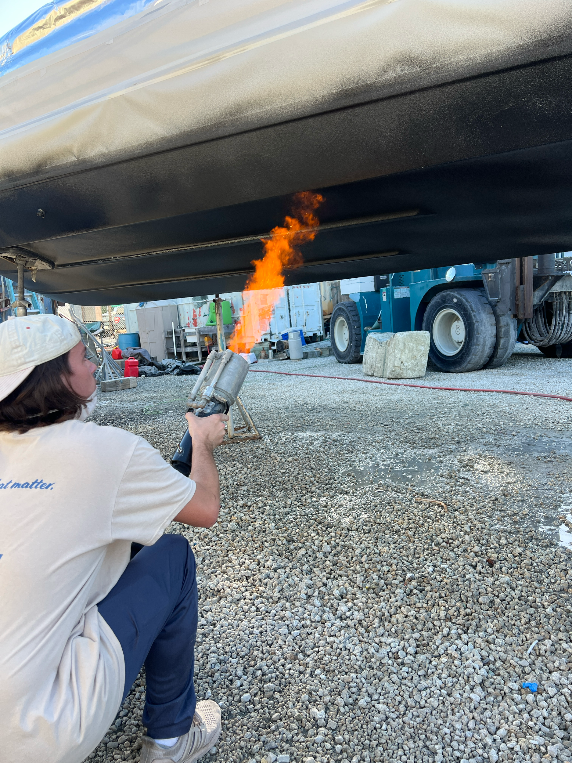 Person safely using a blowtorch to apply heat underneath a boat in an outdoor yard with gravel ground, industrial equipment, and trucks in the background.