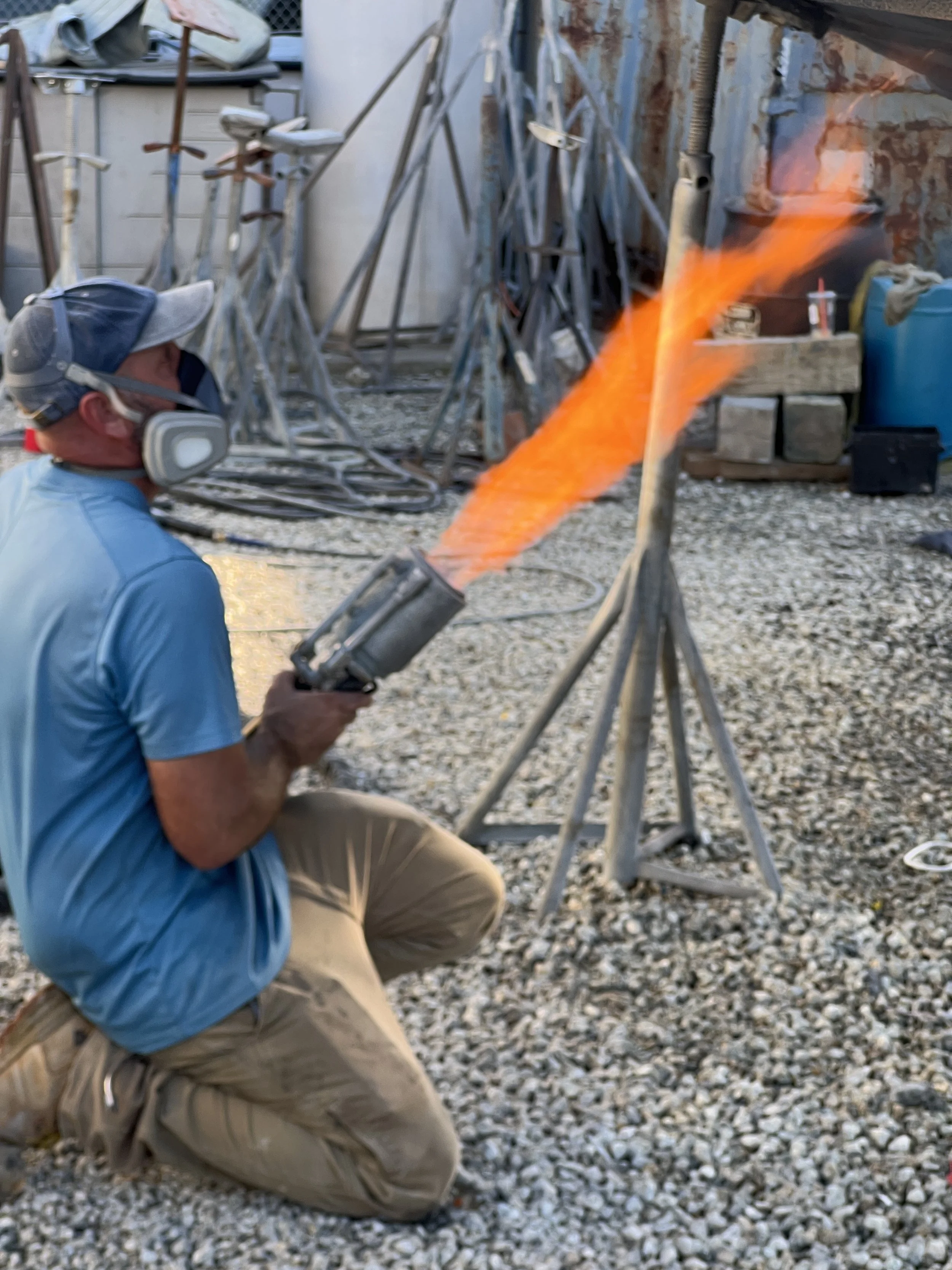 A man kneeling on gravel ground is operating a blowtorch with a large orange flame, wearing a cap and a mask for safety, surrounded by metal equipment and tools in an industrial or workshop setting.