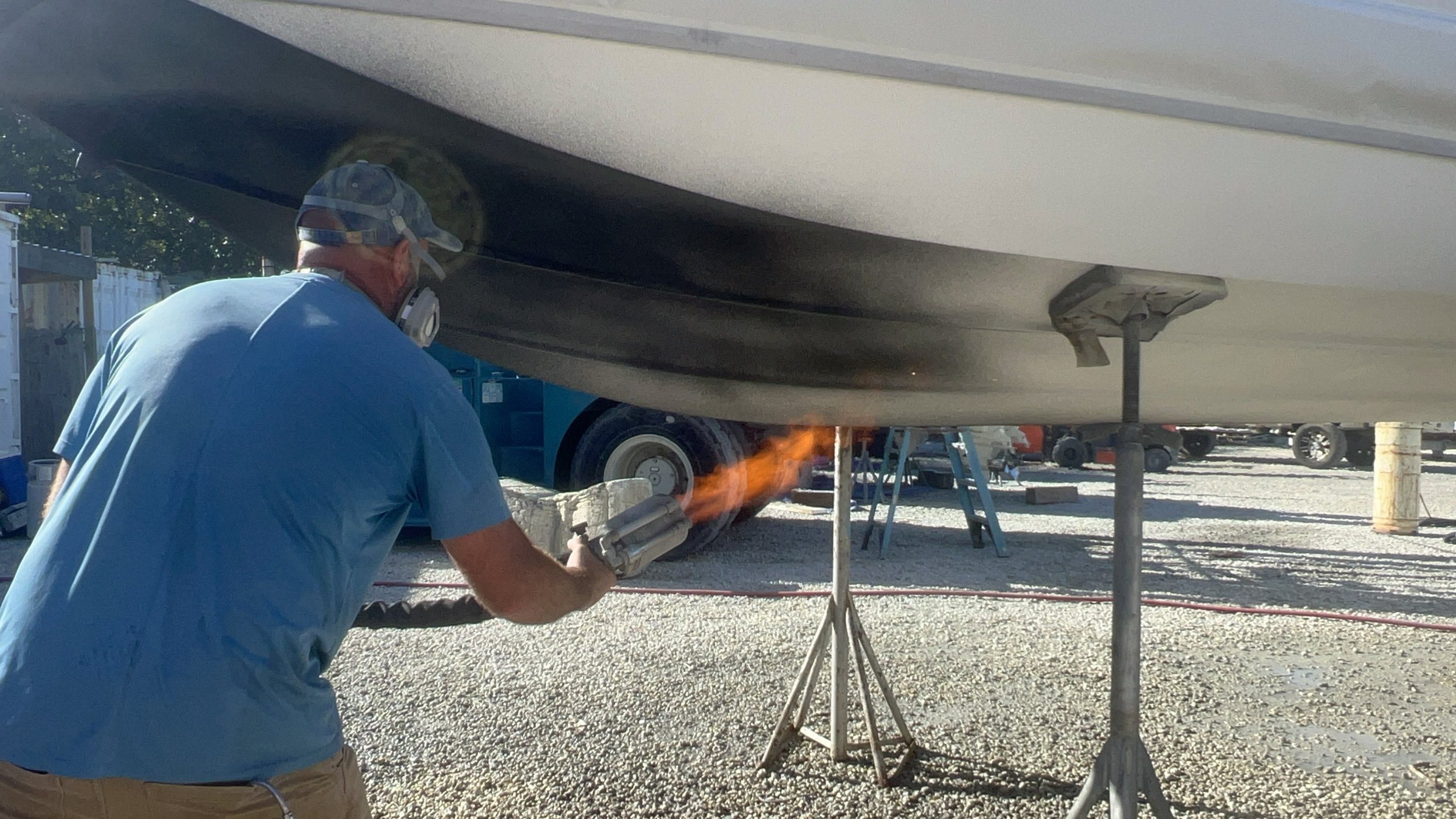 A man working under a boat on land, wearing a blue shirt, cap, and respirator, is welding or grinding, producing sparks and a small flame.