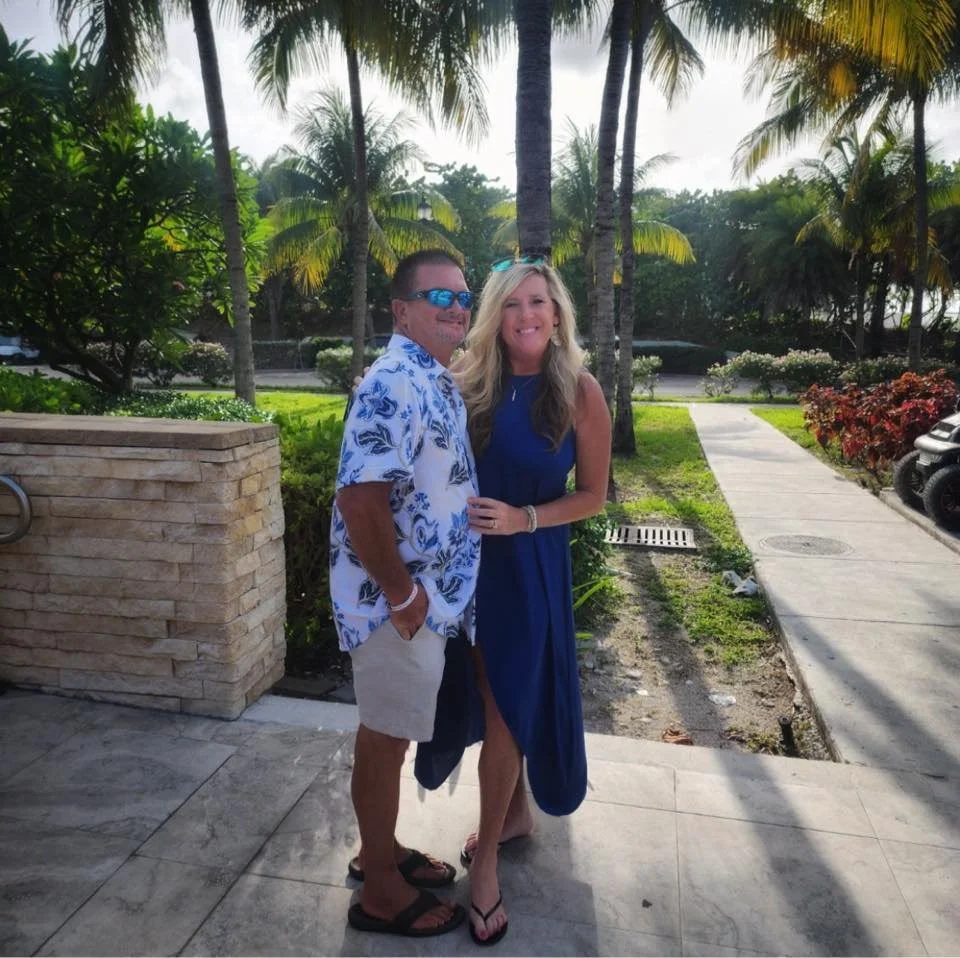 Two people standing on a sidewalk with palm trees in the background, smiling at the camera.
