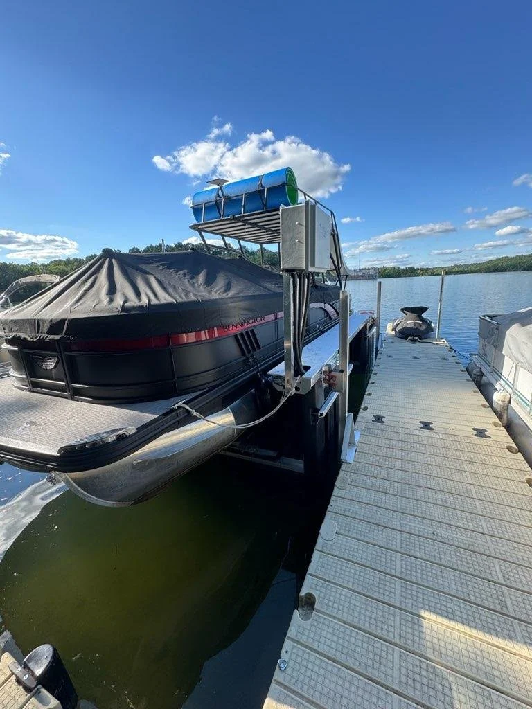 A pontoon boat docked at a marina with a black cover over the boat, floating on a lake under a blue sky with clouds.