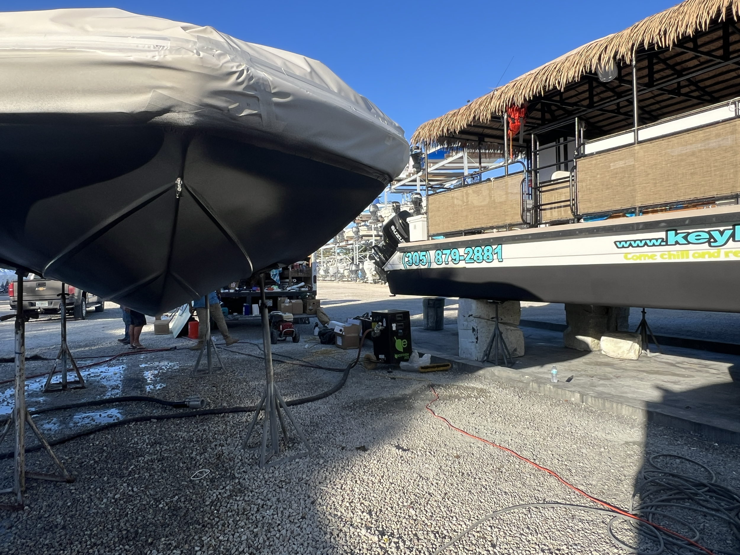 Two boats on land, one covered with a gray tarp and the other with a thatched roof, with equipment and people working nearby, under a clear blue sky.