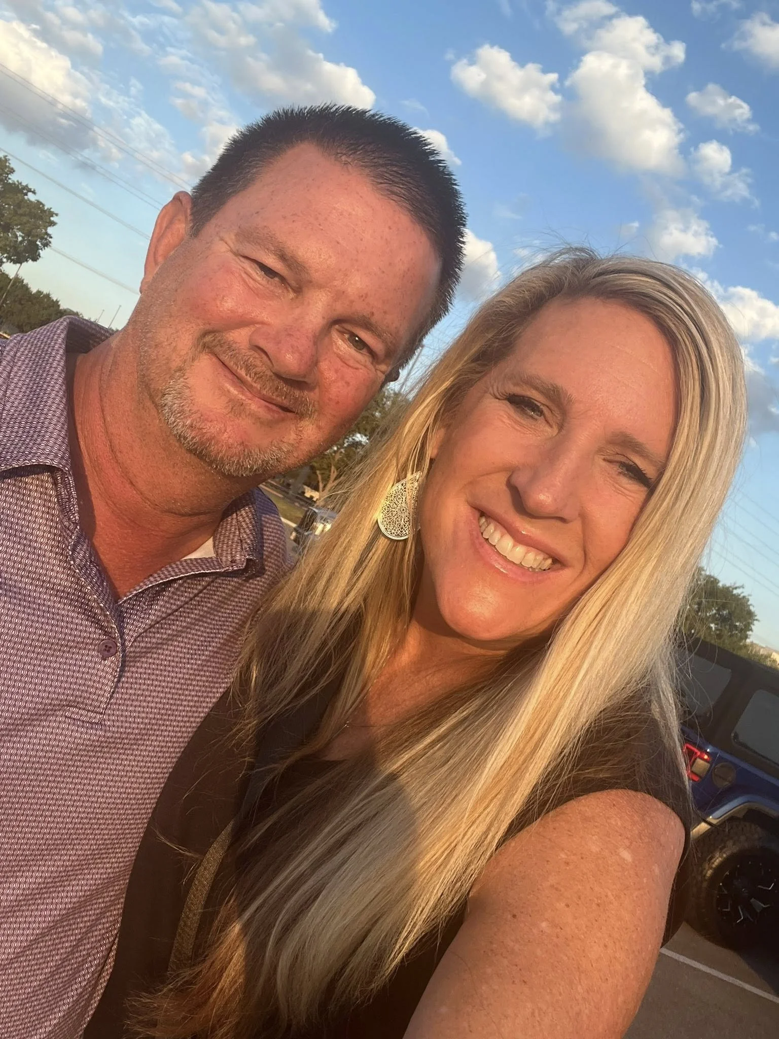 A man and a woman taking a selfie outdoors during sunset, with a partly cloudy sky and parked cars in the background, both smiling.