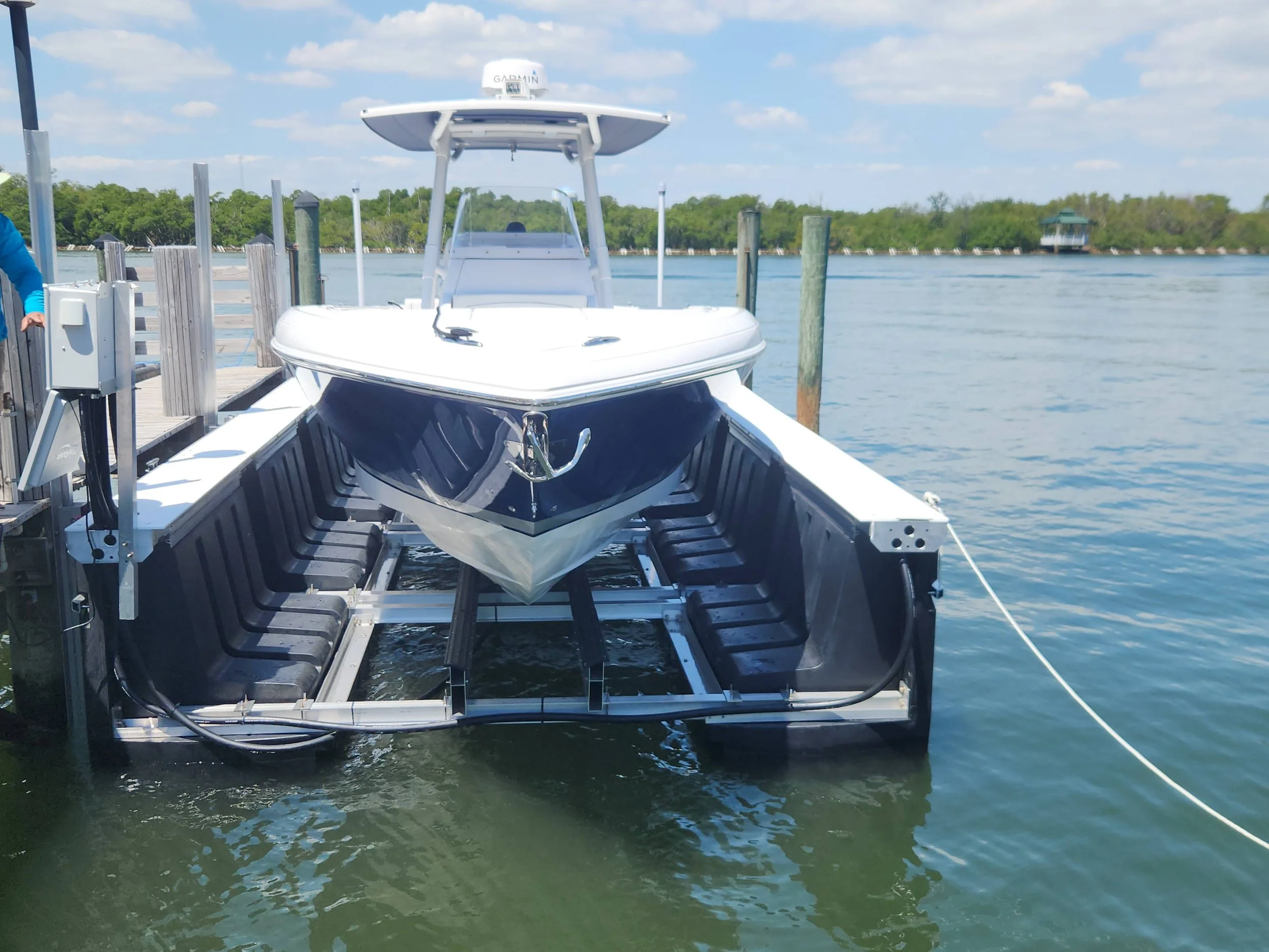 A boat docked at a marina dock with a white and blue boat on a lift, water and trees in the background.