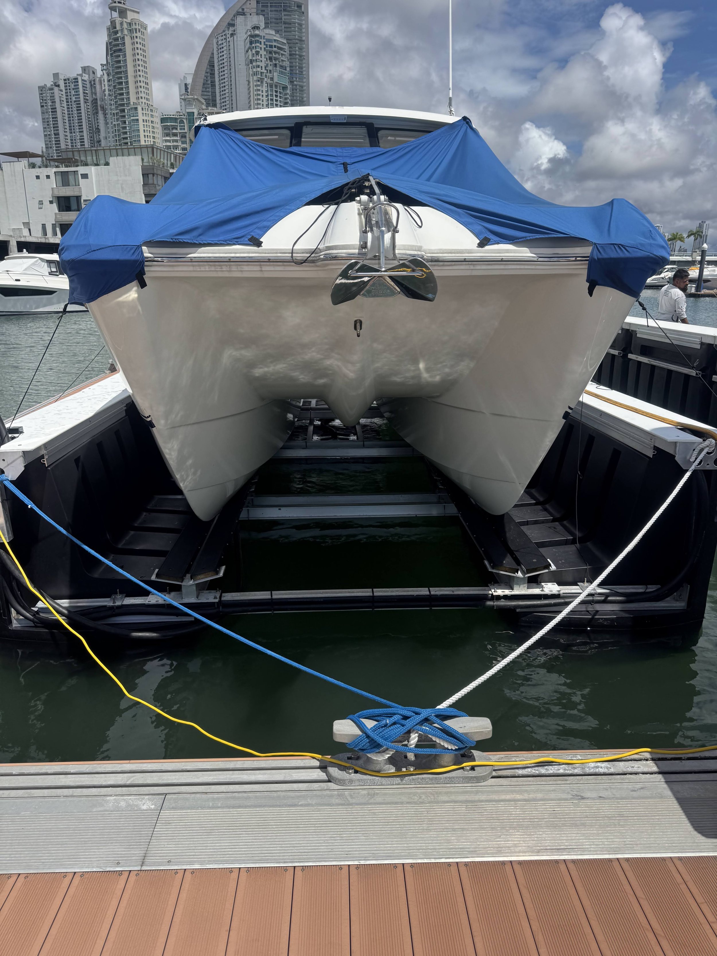 A large white yacht docked at a marina, with a boat cover draped over the front and a city skyline with tall buildings in the background.