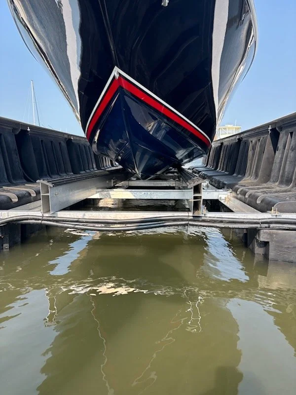 A close-up view of the bow of a large black boat docked in a marina, with the boat on a boat lift and water below reflecting the boat.