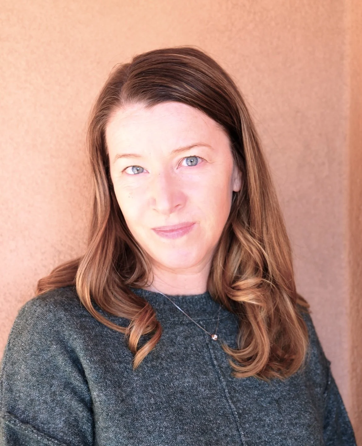 Close-up of a woman with shoulder-length brown hair, blue eyes, wearing a dark gray sweater and a necklace, standing against a textured beige wall.