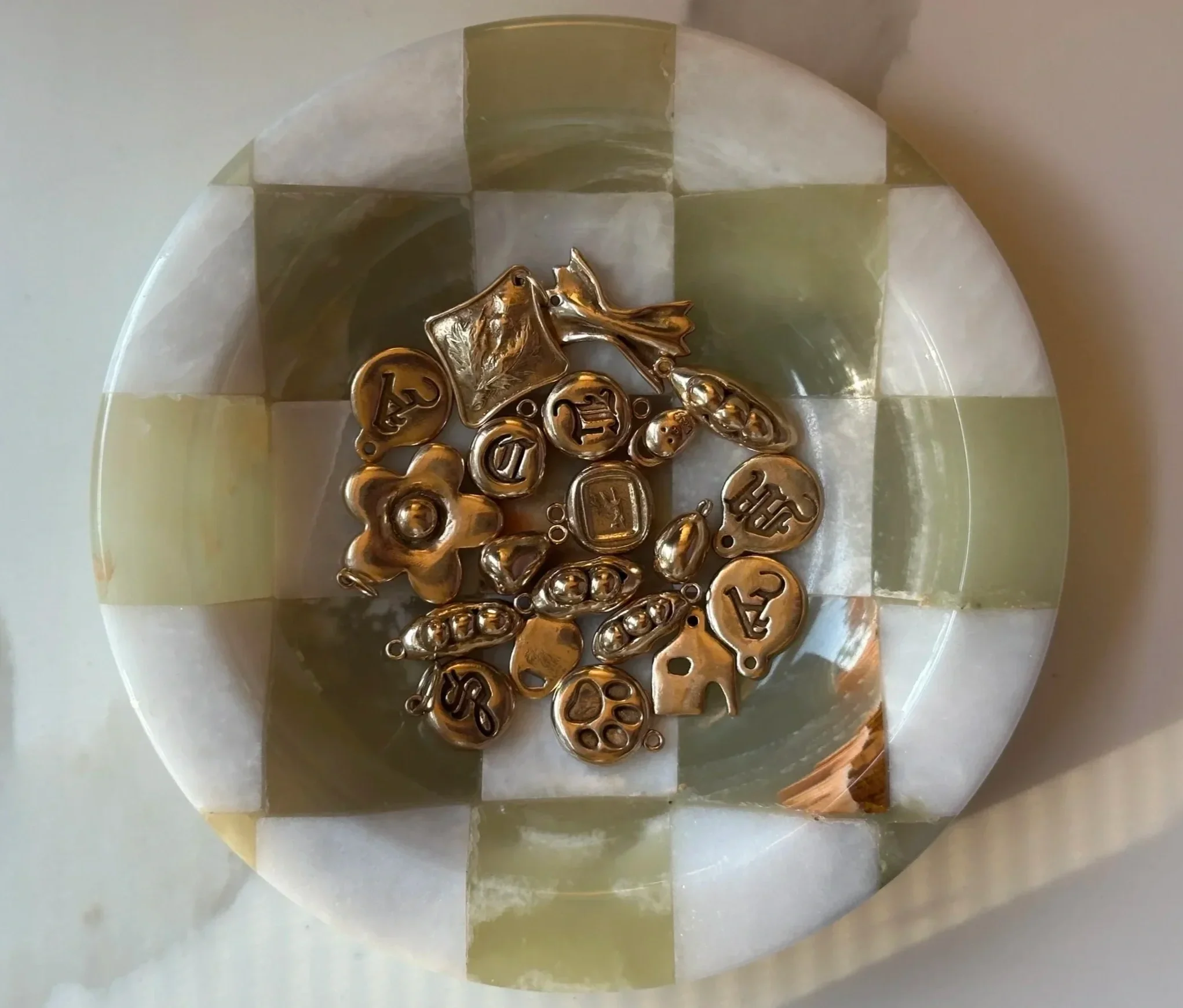 A round mosaic box filled with gold jewelry charms and pendants, placed inside a round mosaic container with a checkered pattern of green and beige tiles.