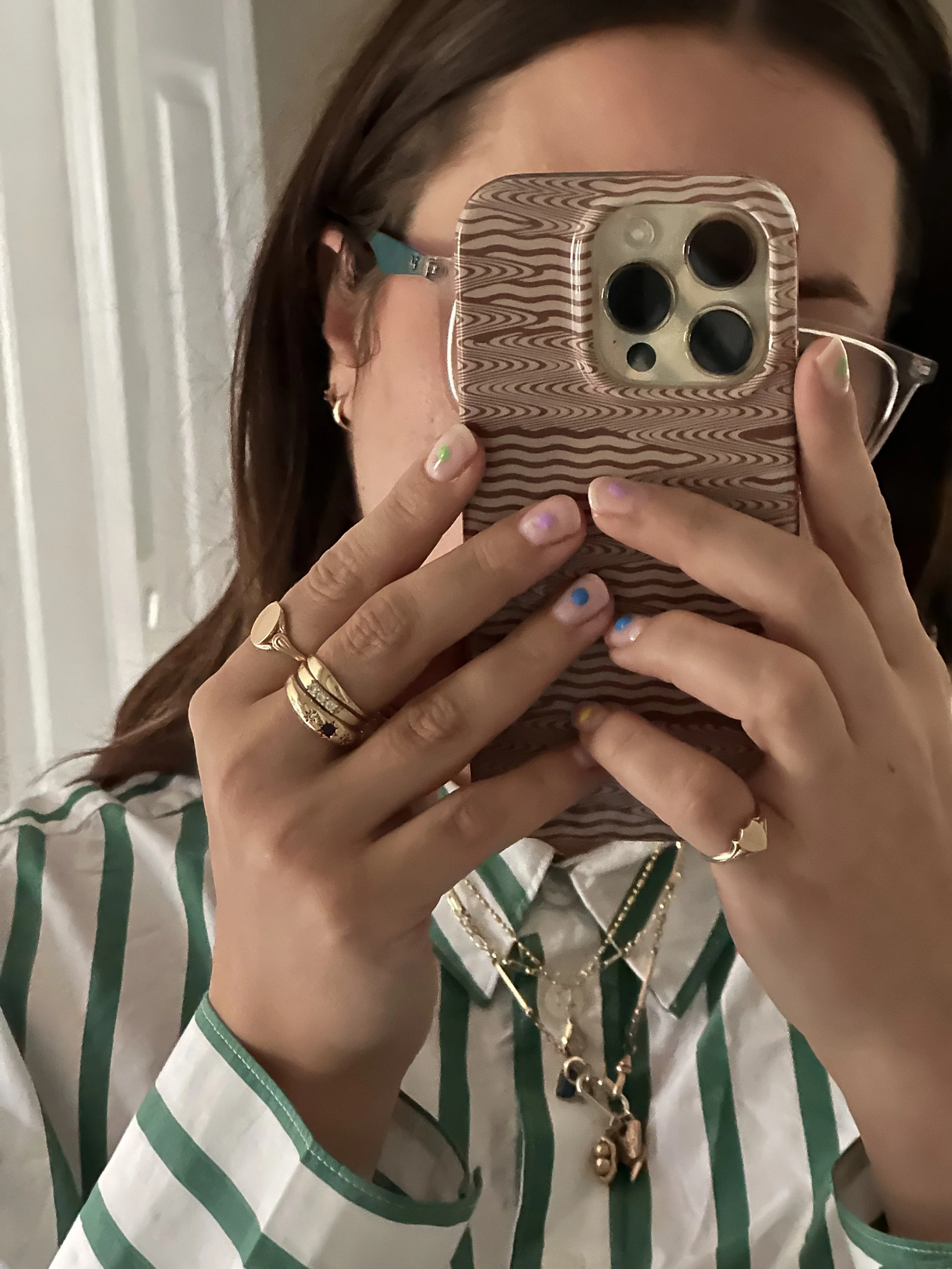 A woman taking a mirror selfie with her phone, showing her face partially covered by the phone. She has shoulder-length dark hair, wears glasses with teal frames, and is dressed in a white and green striped shirt. She has various rings on her fingers, a layered gold necklace, and colorful nail polish.