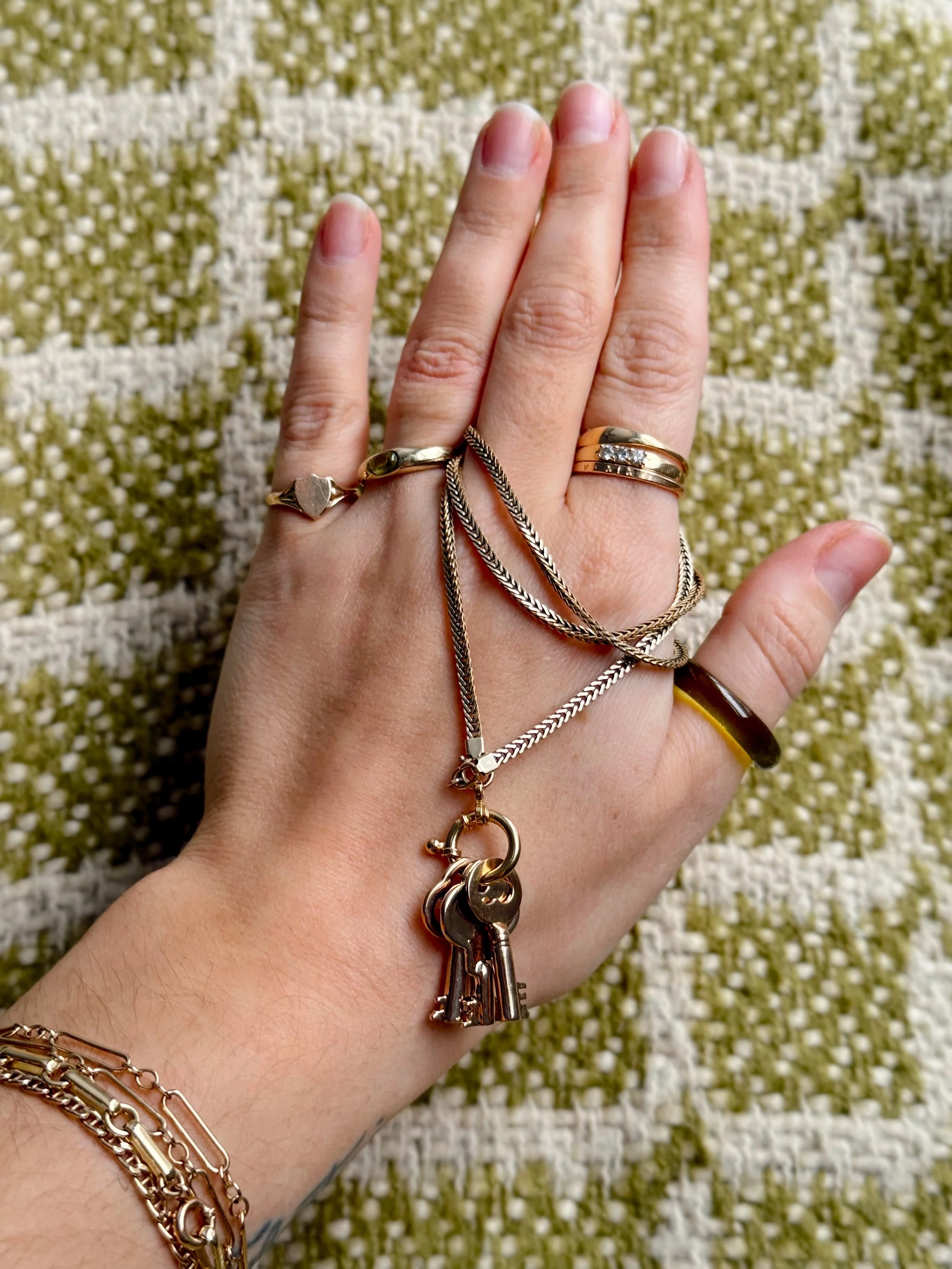 A hand wearing multiple rings and bracelets, holding several necklaces with key pendants, resting on a textured, patterned surface.