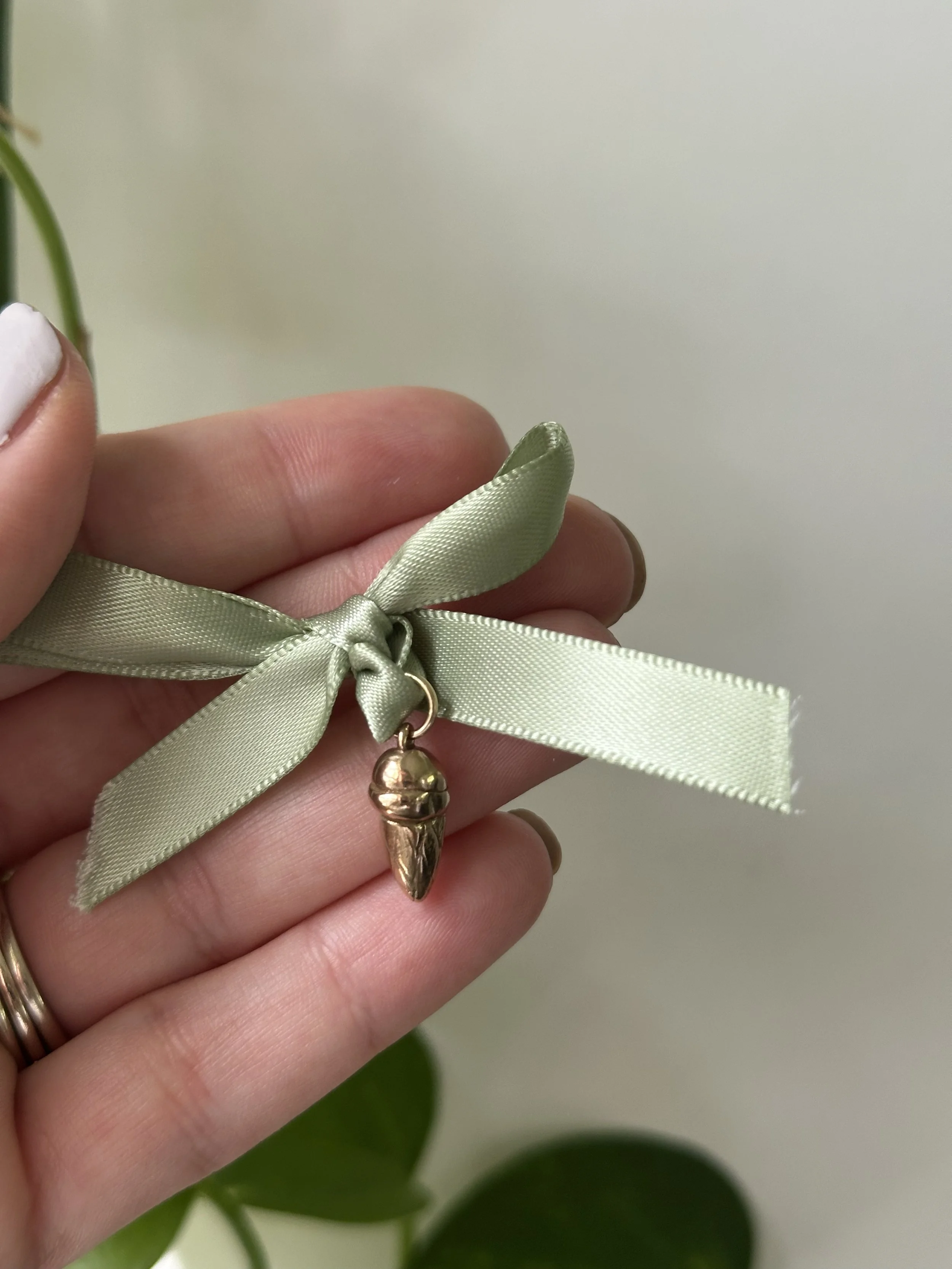 A hand holding a green ribbon tied in a bow with a small bronze ice cream cone charm hanging from it, against a plain light background.