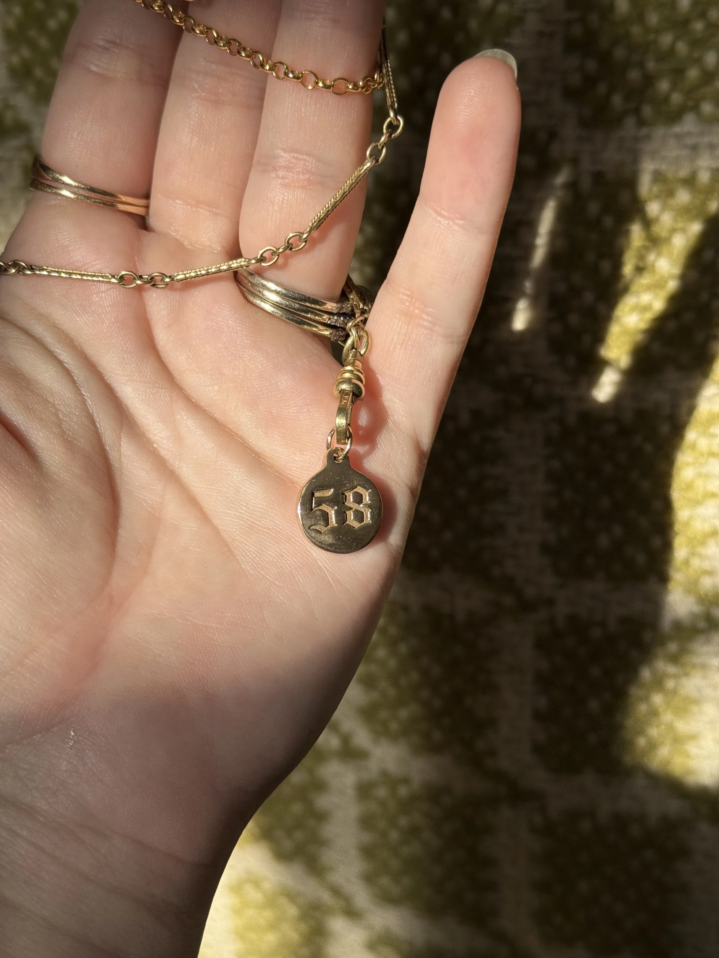 A hand holding multiple gold necklaces with a small round pendant engraved with the number 58.