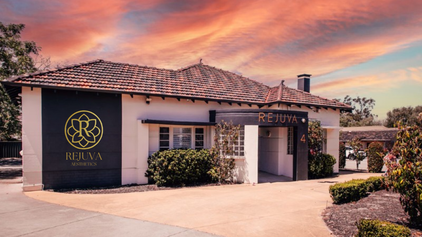 Exterior of a building with the name 'REJUVA' and the word 'AESTHETICS' on the side, surrounded by landscaped bushes and trees, at sunset with a colorful sky.