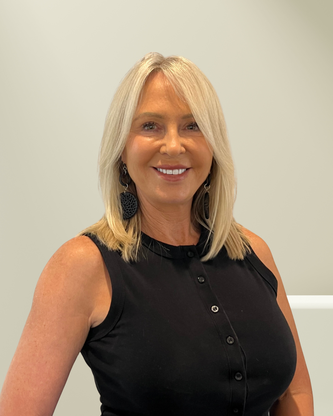 A smiling woman with shoulder-length black hair wearing a blue top, standing outdoors with green foliage in the background.