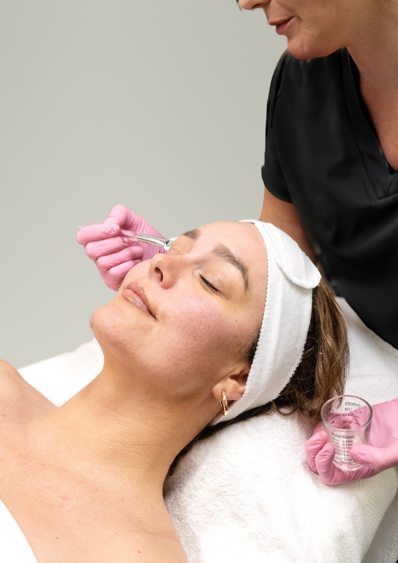 A woman receives a facial treatment from a professional wearing pink gloves in a clinical setting.