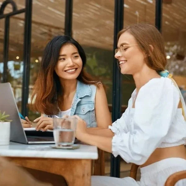 Two women sitting at a table, smiling and chatting, with a laptop and a glass of water in front of them, in a cozy coffee shop or cafe.