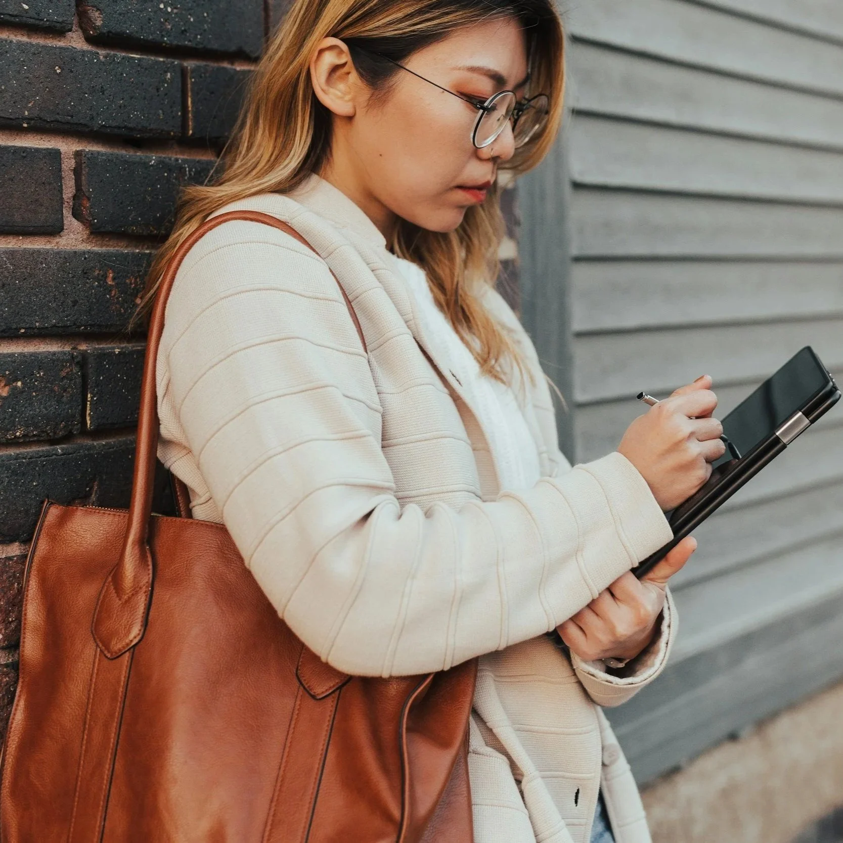 A woman wearing glasses, a beige jacket, and carrying a large brown leather bag, is standing against a brick wall and looking at her phone.