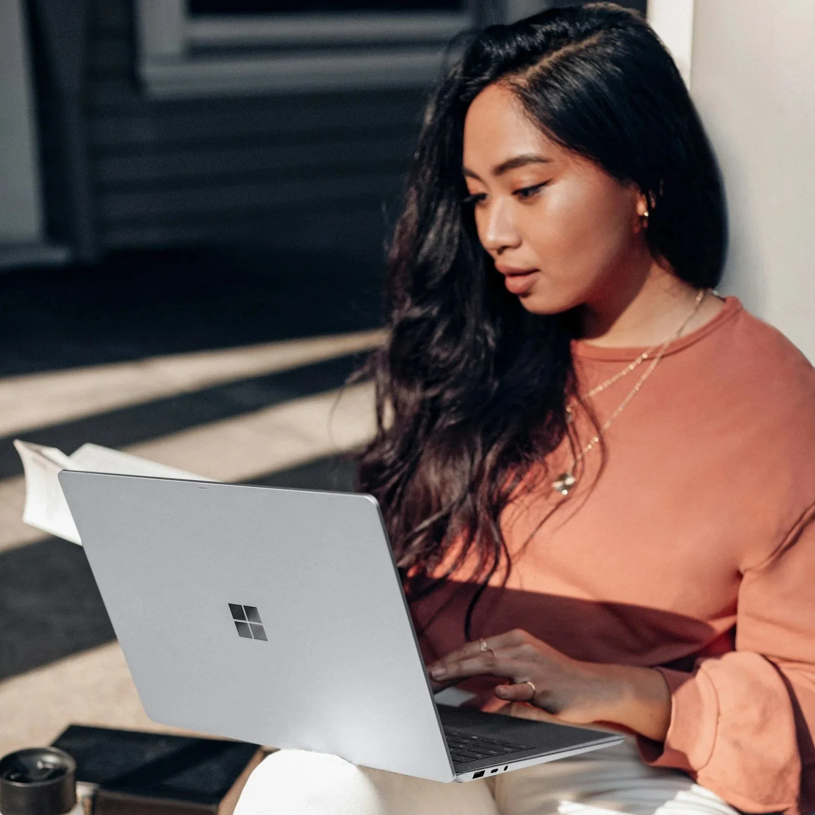 A young woman with long dark hair, wearing a peach-colored top and jewelry, sitting outdoors and working on a silver Microsoft laptop.