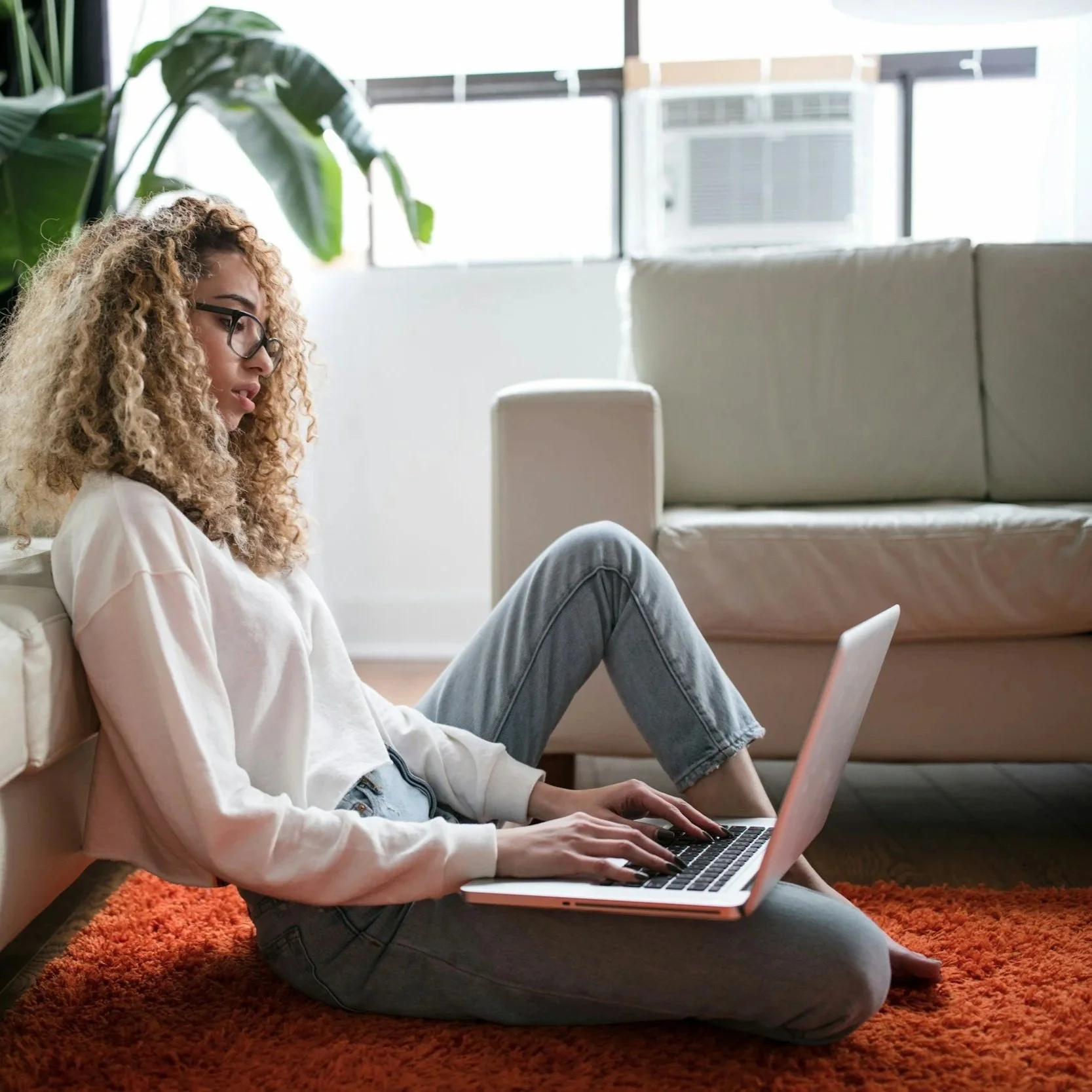 A young woman with curly hair and glasses sitting on an orange rug, leaning against a beige sofa and working on a laptop in a bright living room with large windows.