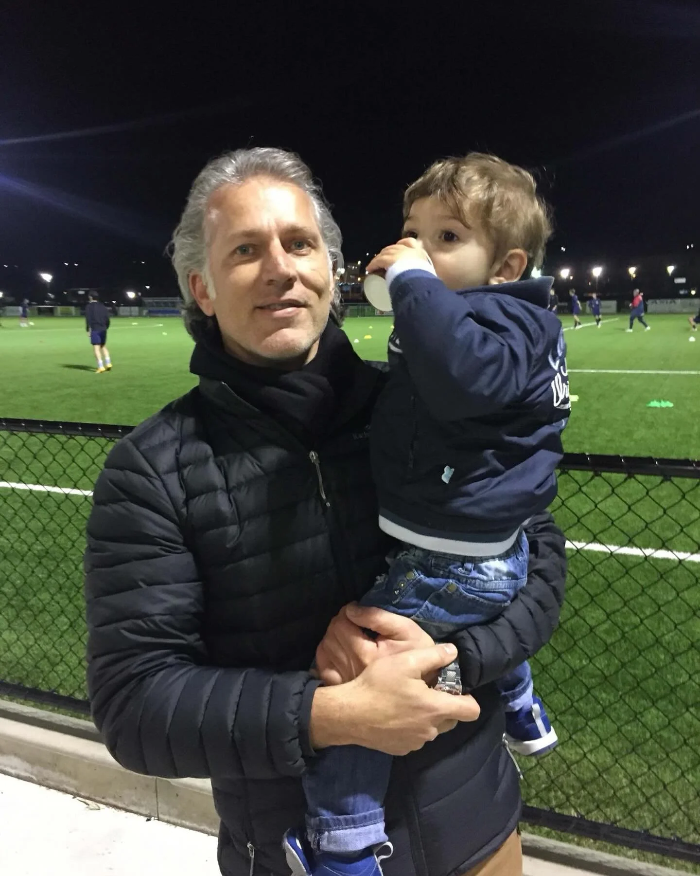 Man holding a child at a soccer field at night, with players practicing in the background.