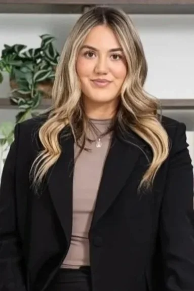 A young woman with blonde hair wearing a black blazer and gray top, sitting in an office setting with a plant and bookshelf in the background.