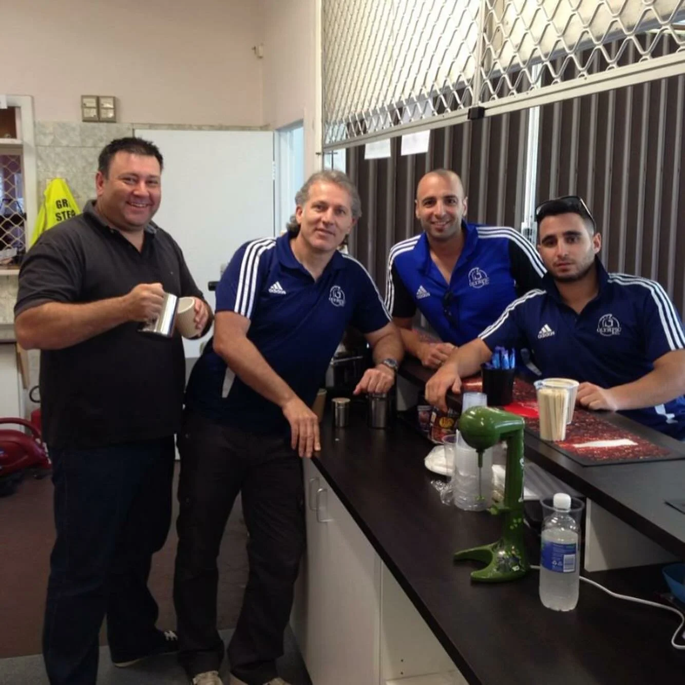 Four men in blue sports shirts with white stripes, inside a room with a counter and barred window, standing and sitting at the counter with drinks and condiments, smiling at the camera.