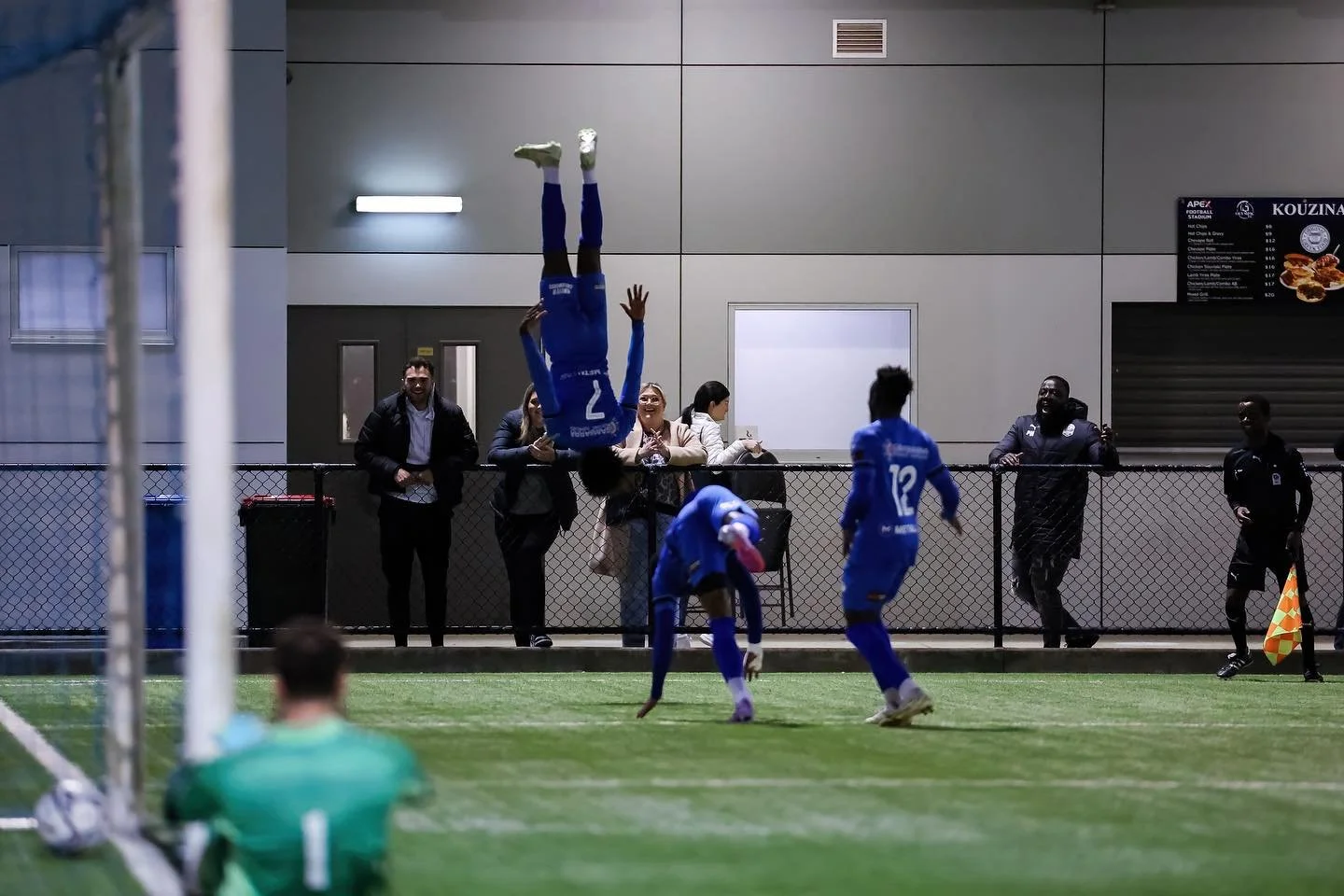 Soccer players celebrating a goal, with one player performing a headstand on the field, spectators and officials watching from behind a fence.