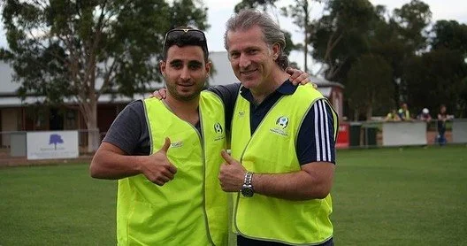 Two men wearing yellow safety vests standing on a sports field, smiling and giving thumbs up.