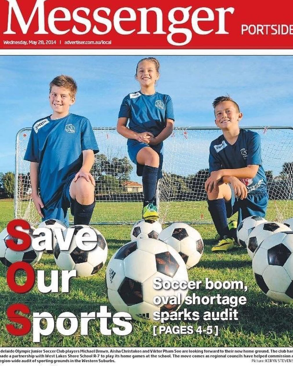 Three young children wearing blue soccer uniforms are on a soccer field in front of a goal. They are surrounded by soccer balls and are smiling at the camera.