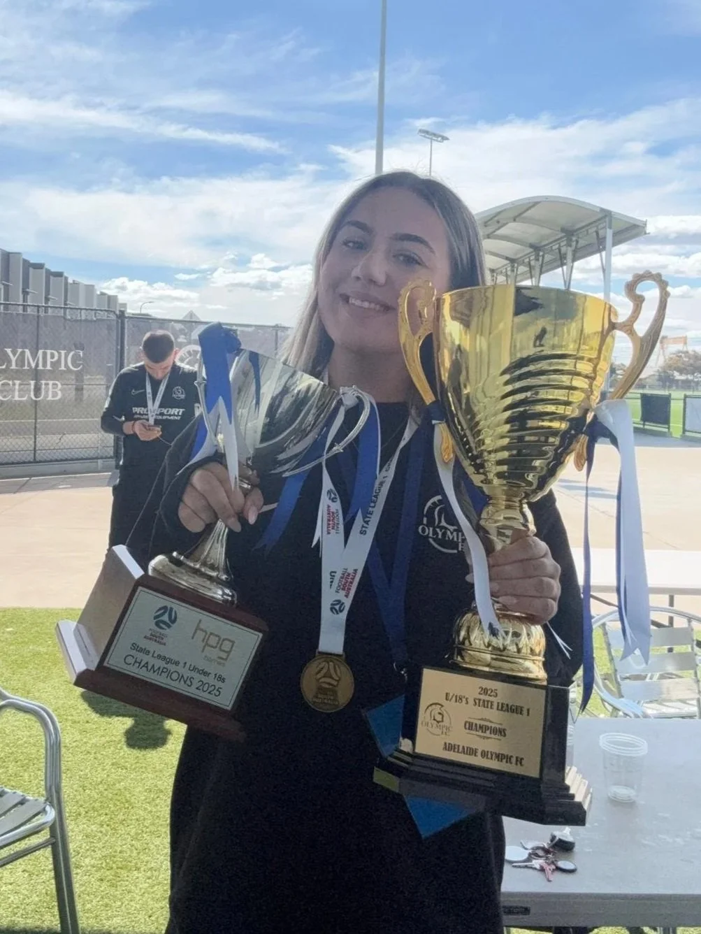 A young woman smiling and holding two large trophies on a sports field, with medals around her neck. One trophy reads '2025 A/18'S STATE LEAGUE I CHAMPIONS ADELAIDE OLYMPIC FC,' and the other reads 'HPA State League 1 Under 18 CHAMPIONS 2025.' A person in the background is looking at their phone.