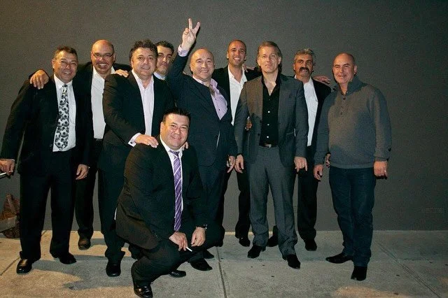 Group of ten men dressed in formal and semi-formal attire, standing outdoors against a dark wall, with some smiling and one man making a peace sign.