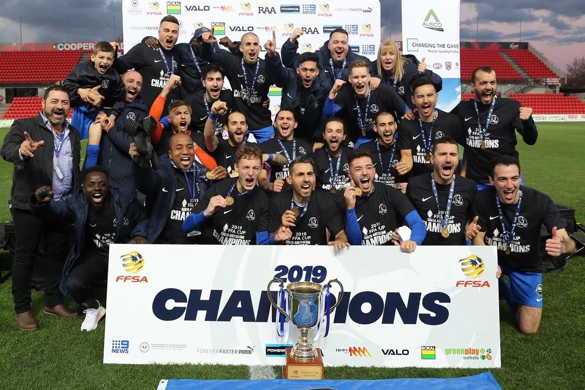 A group of soccer players and coaches celebrating their victory at a stadium, holding medals, posing behind a large sign that reads "2019 Champions." A trophy is in front of the sign, and the group is smiling and raising their fists in celebration.
