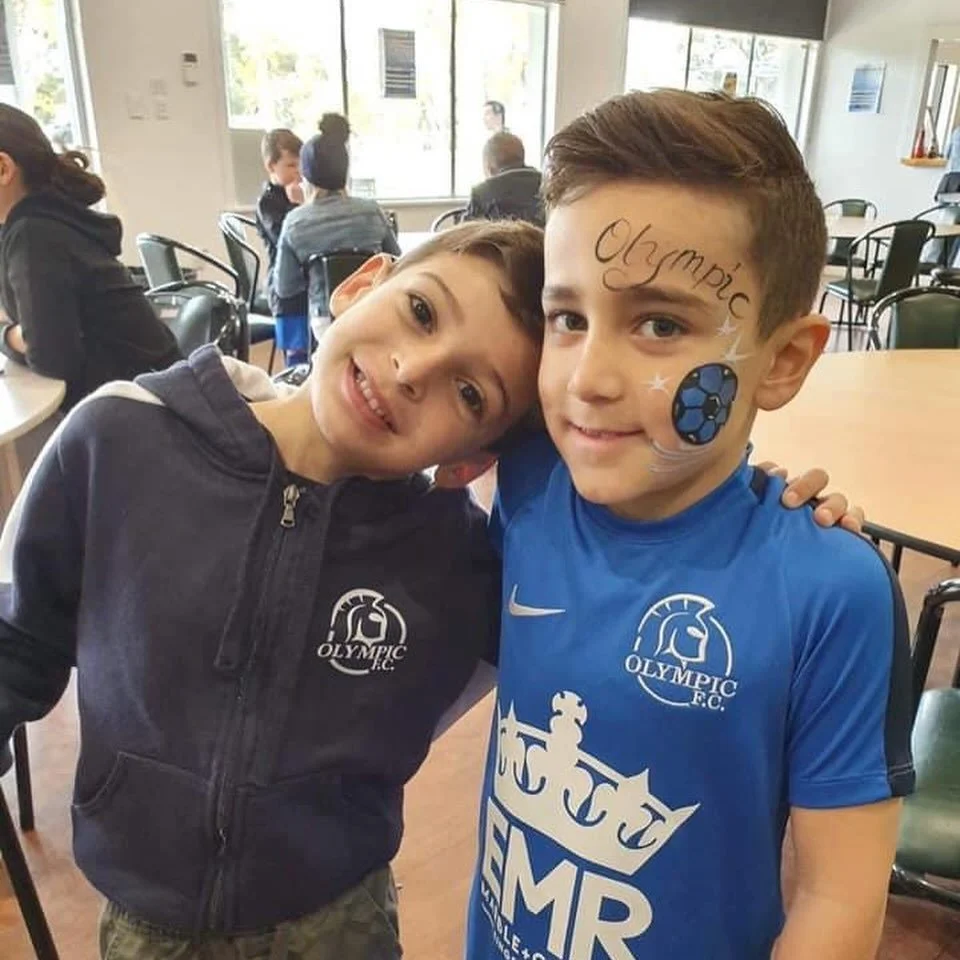 Two young boys at a sports event with face paint, wearing team gear, smiling and posing together inside a room with tables and other children in the background.