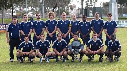 Group of eleven men in soccer uniforms on a grassy field, posing for a team photo.
