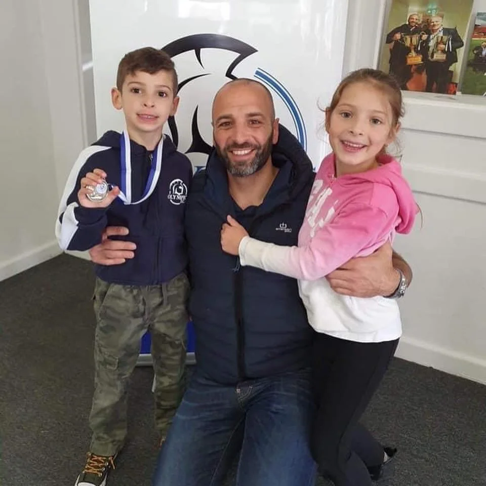 A man kneeling down surrounded by a boy and a girl, all smiling warmly. The boy is holding a silver medal, and the girl is embracing the man. They are in an indoor setting with a logo and photos visible on the wall behind them.
