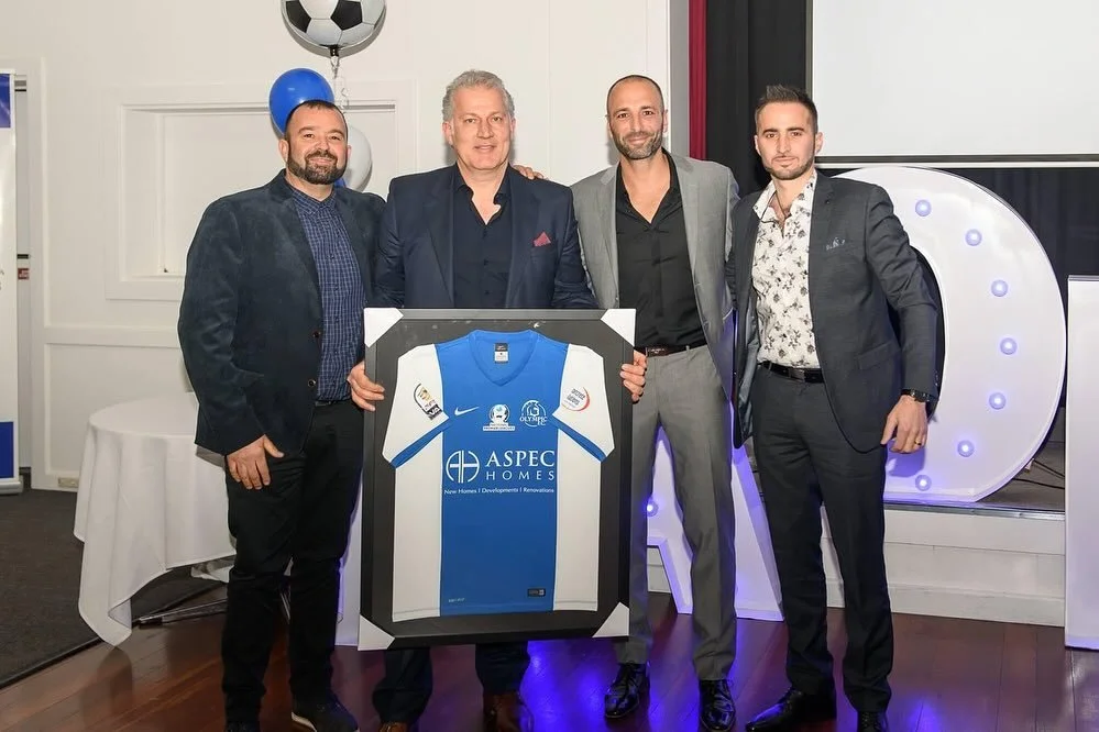 Four men in suits holding a framed soccer jersey, standing in a decorated event space with balloons and a large illuminated letter D behind them.