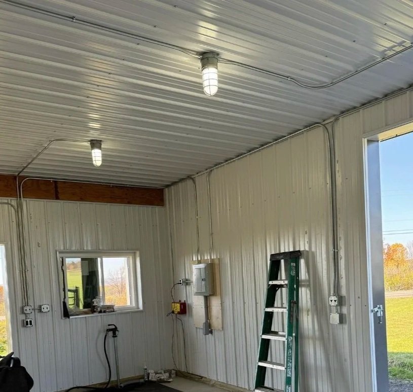 The image shows the interior of a metal building with a corrugated metal ceiling and wall panels, illuminated by two ceiling lights. There is a small window and a door open to outside green fields, with some trees and power lines visible in the distance. Various electrical outlets, boxes, a green ladder, and a black hose are also visible inside.