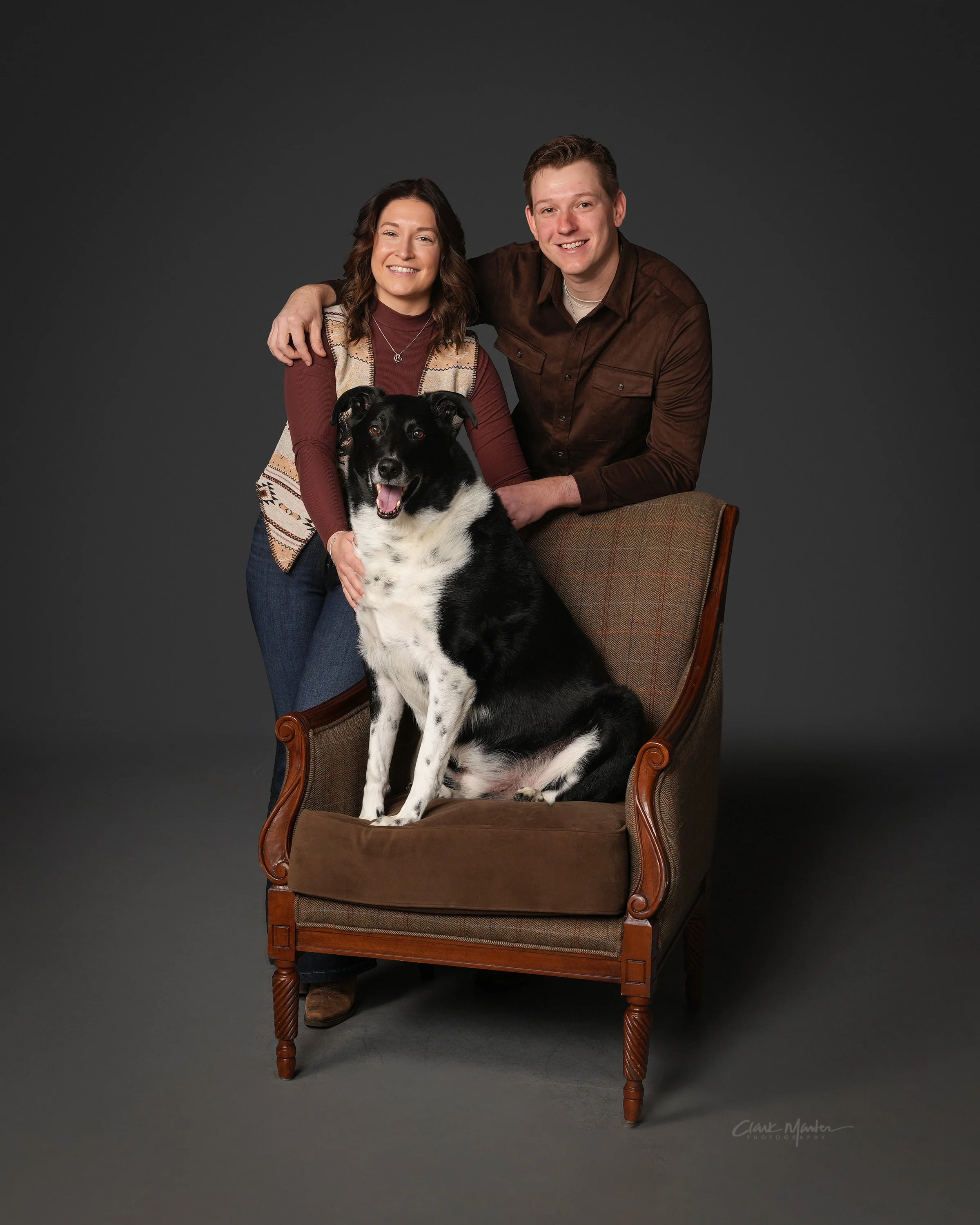 A happy couple sitting together on a vintage wooden armchair, posing with their black and white coated dog against a dark grey background. The woman has shoulder-length wavy brown hair and is wearing a patterned vest over a maroon long-sleeve top. The man, with short brown hair, is dressed in a brown button-up shirt. They are smiling, and the dog is sitting on the chair with its tongue out, looking directly at the camera.