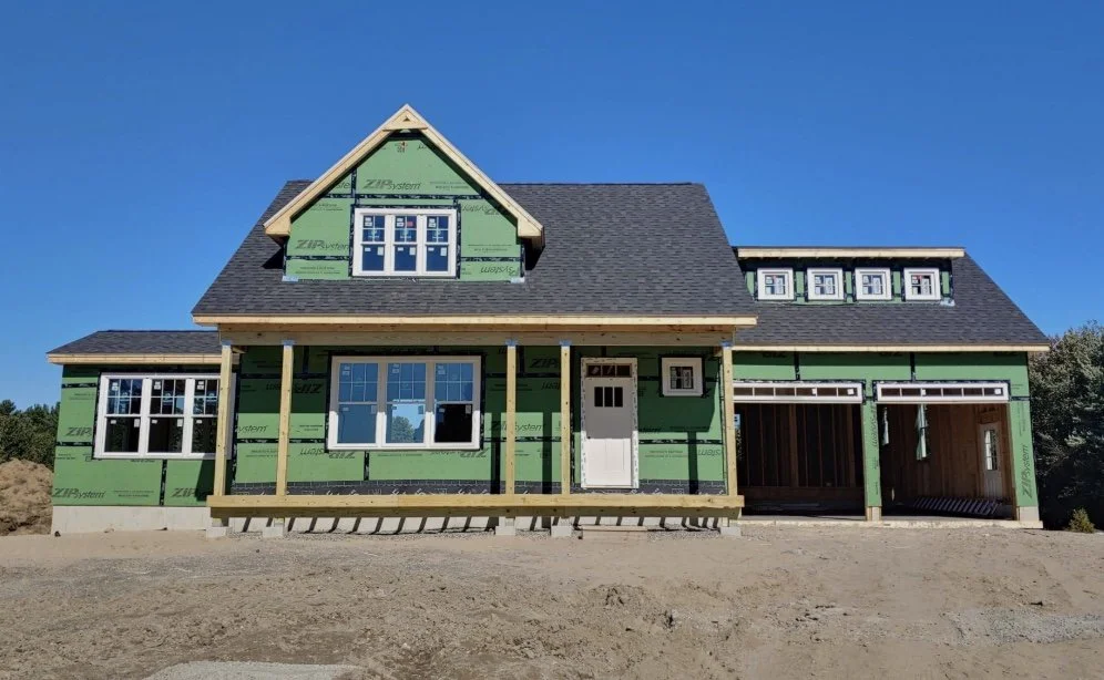 Newly constructed house with green sheathing and unfinished exterior, featuring multiple windows, a covered front porch, and a two-car garage, under a clear blue sky.