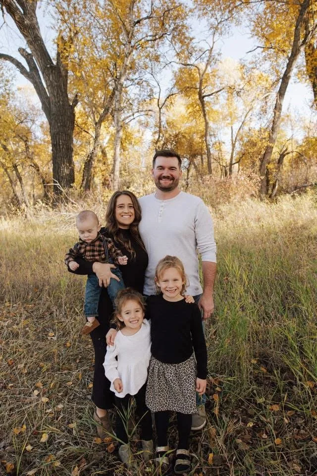 A family of five outdoors in a field with fall-colored trees in the background. The parents are smiling, with the mother holding a young child, and two girls standing in front of them.