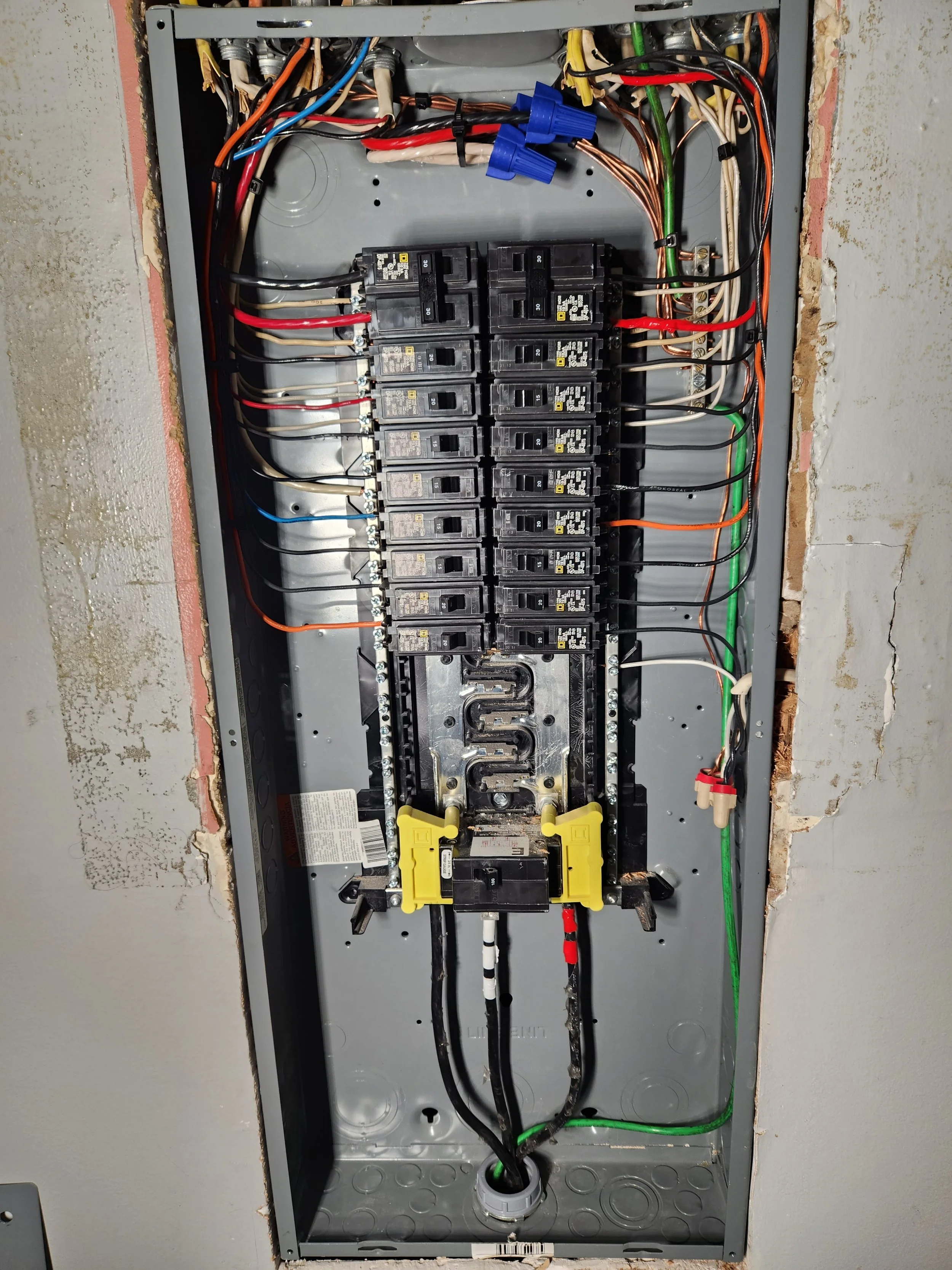 An open electrical panel with multiple black circuit breakers, various colored wires, and green grounding wire, mounted on a wall with exposed drywall edges.