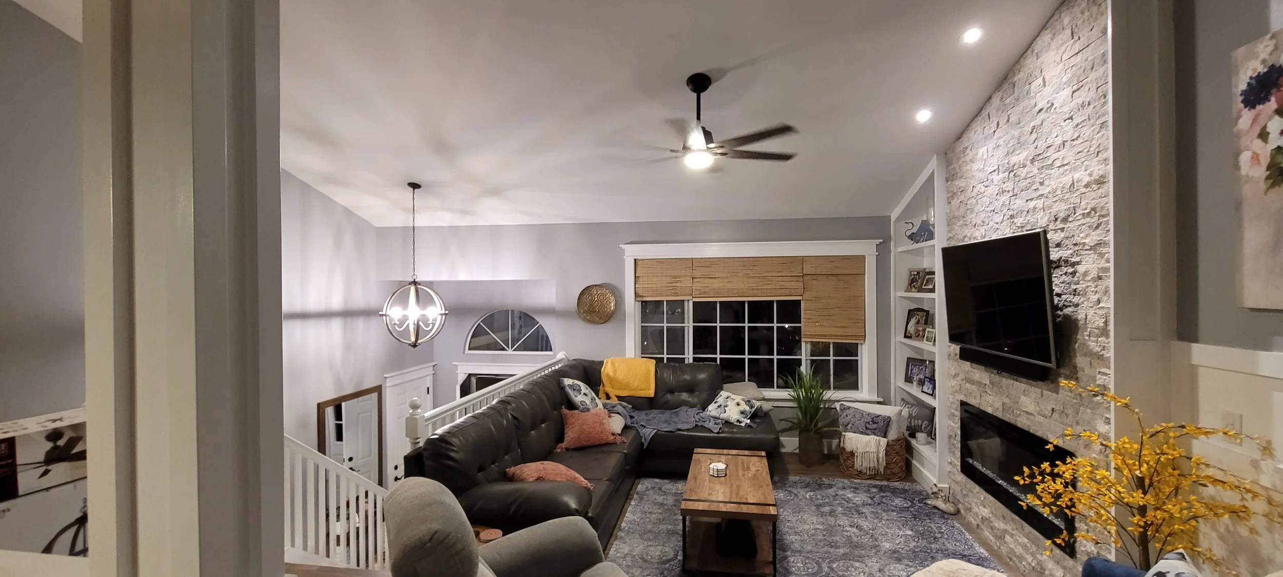 A cozy living room with a black sectional sofa, a gray armchair, a wooden coffee table, and a stone fireplace with a mounted TV. There's a ceiling fan and a decorative chandelier, windows with bamboo blinds, and shelves with framed photos and decor.