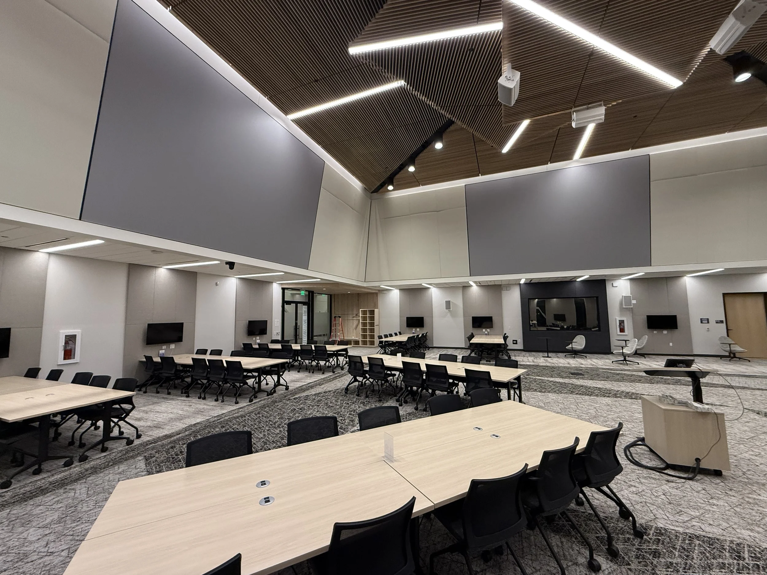 A modern conference or meeting room with multiple long tables and black chairs. The ceiling has wooden panels with integrated linear lighting and speakers. The walls are light-colored with mounted screens and a black fireplace or media unit.