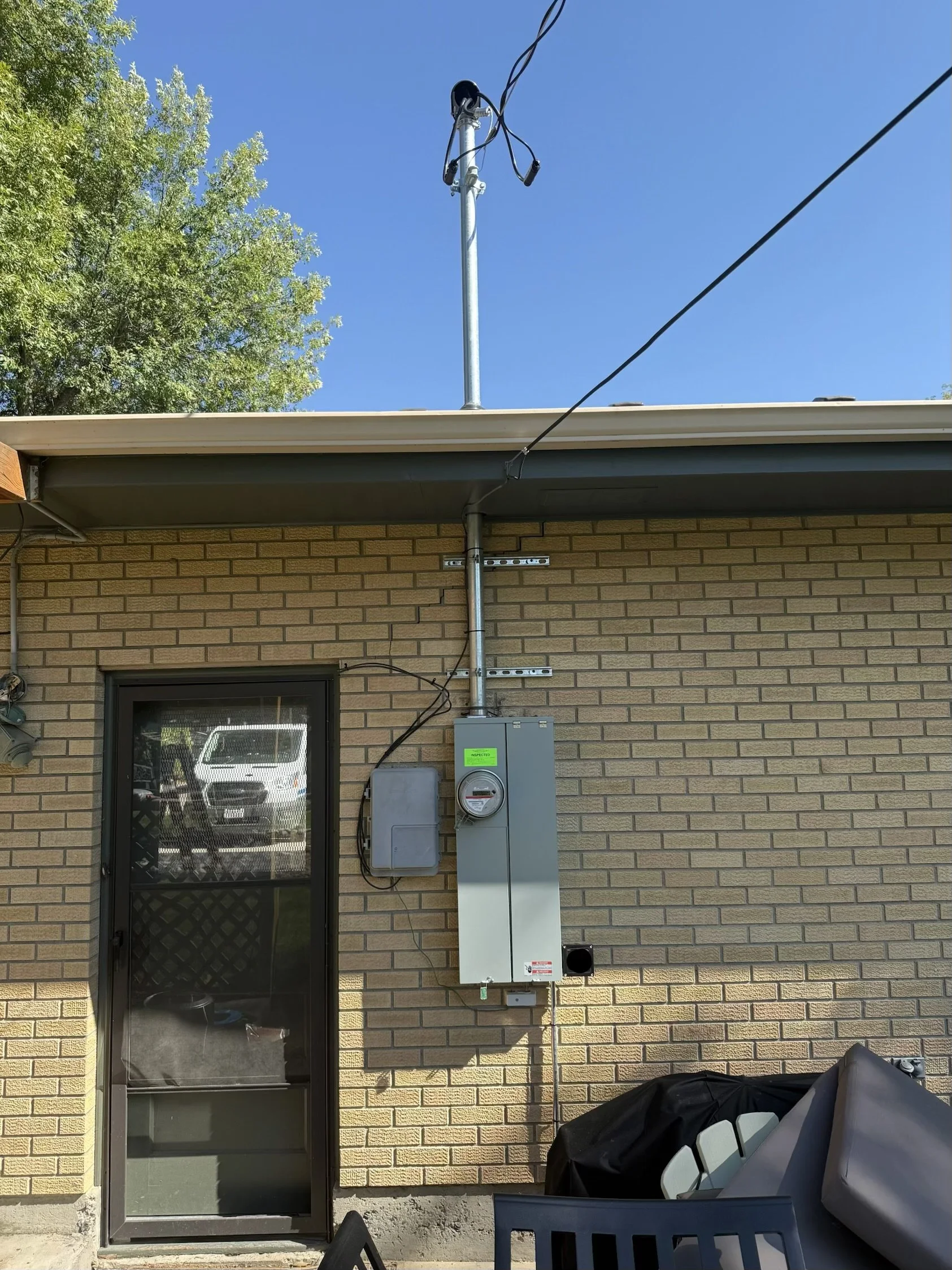 Back view of a brick house wall with electrical panel and meter, a sliding glass door, and outdoor furniture covered with a tarp. There is a tree and clear blue sky overhead.