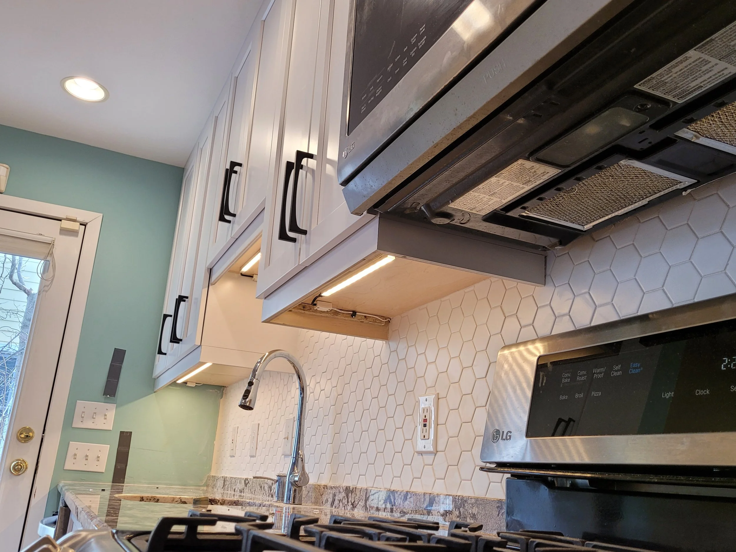 Modern kitchen with white cabinets, a marble island, a black cooktop, a metallic range hood, and a white honeycomb backsplash, under a large window and recessed ceiling lights.
