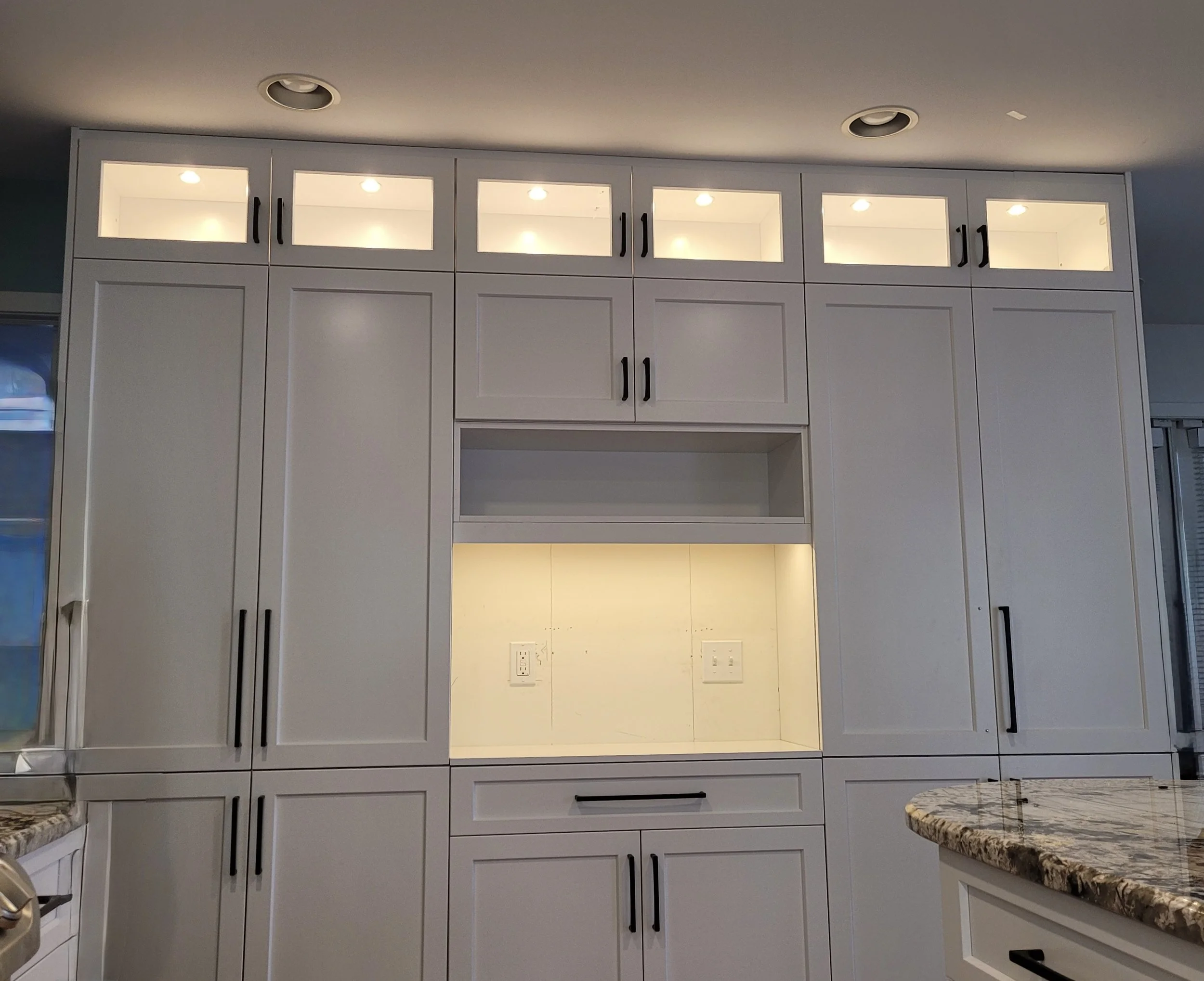 Kitchen upper cabinets with glass doors, black handles, and lighting inside, above a granite countertop. Part of a kitchen with white walls and electrical outlets visible.