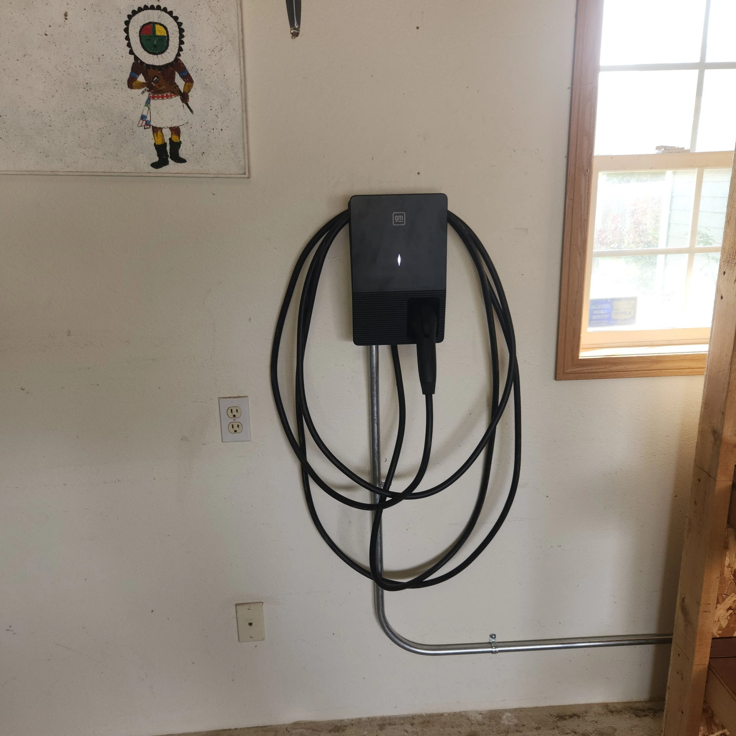 A wall with a painted picture of a Native American figure, an electrical outlet, and a black electronic device with cables. There is a window with a wooden frame and sunlight coming through on the right side.