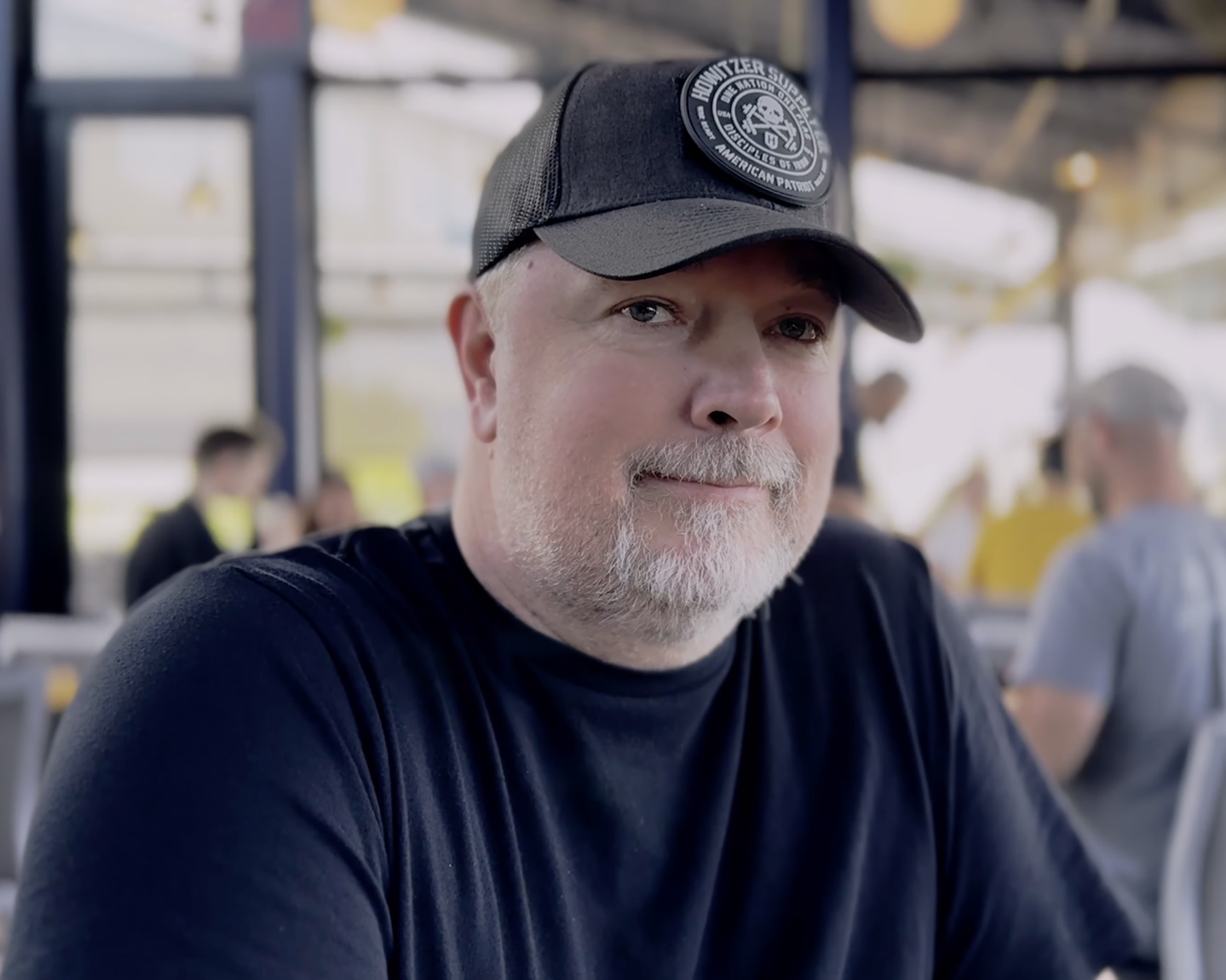 A man with a gray beard wearing a black cap with a patch that reads 'Howitzer Studio' and a black t-shirt, sitting in a busy outdoor setting.