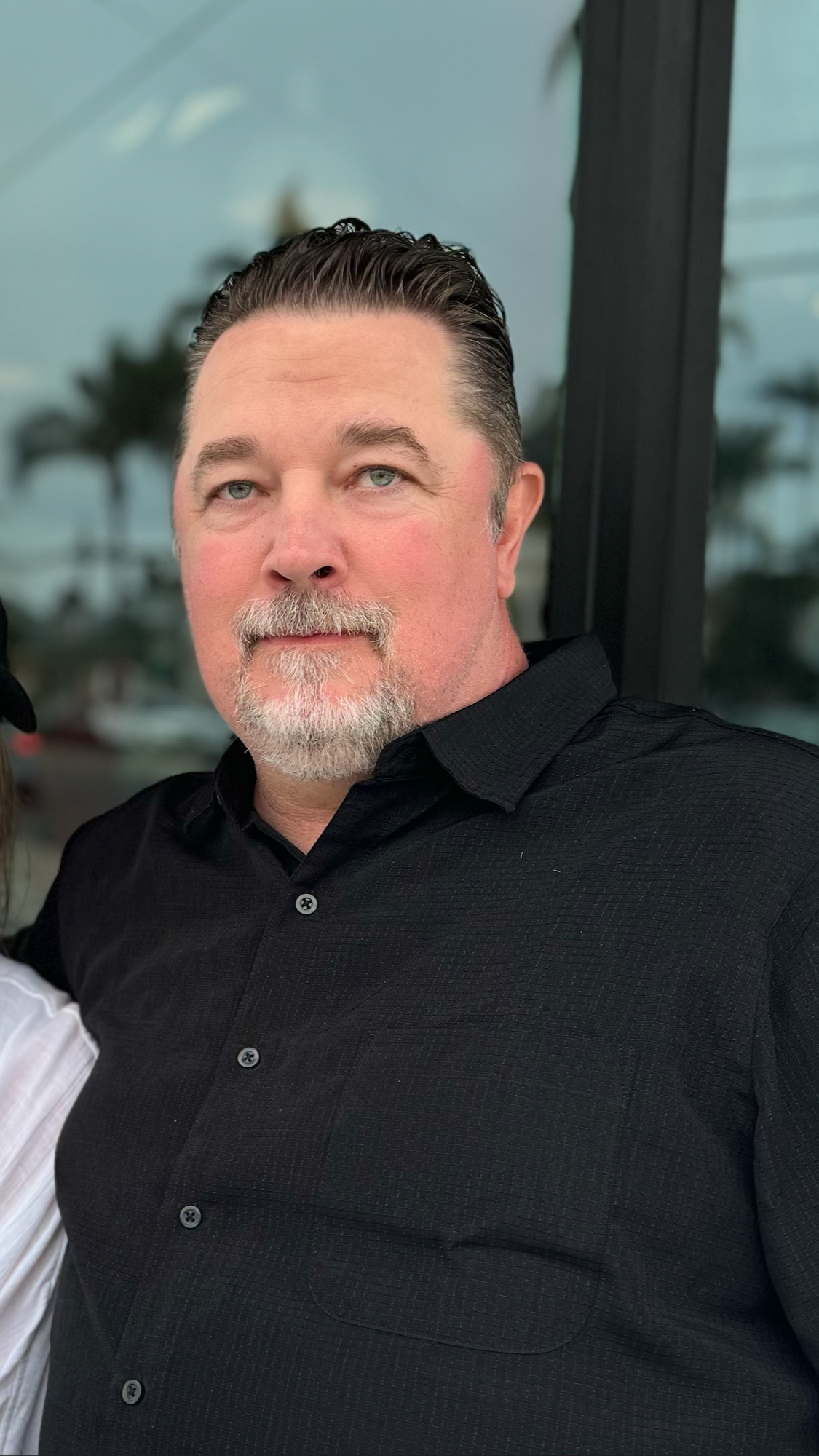 A middle-aged man with short dark hair and a gray beard, wearing a black collared shirt, stands outdoors in front of glass with a blurred background of palm trees.